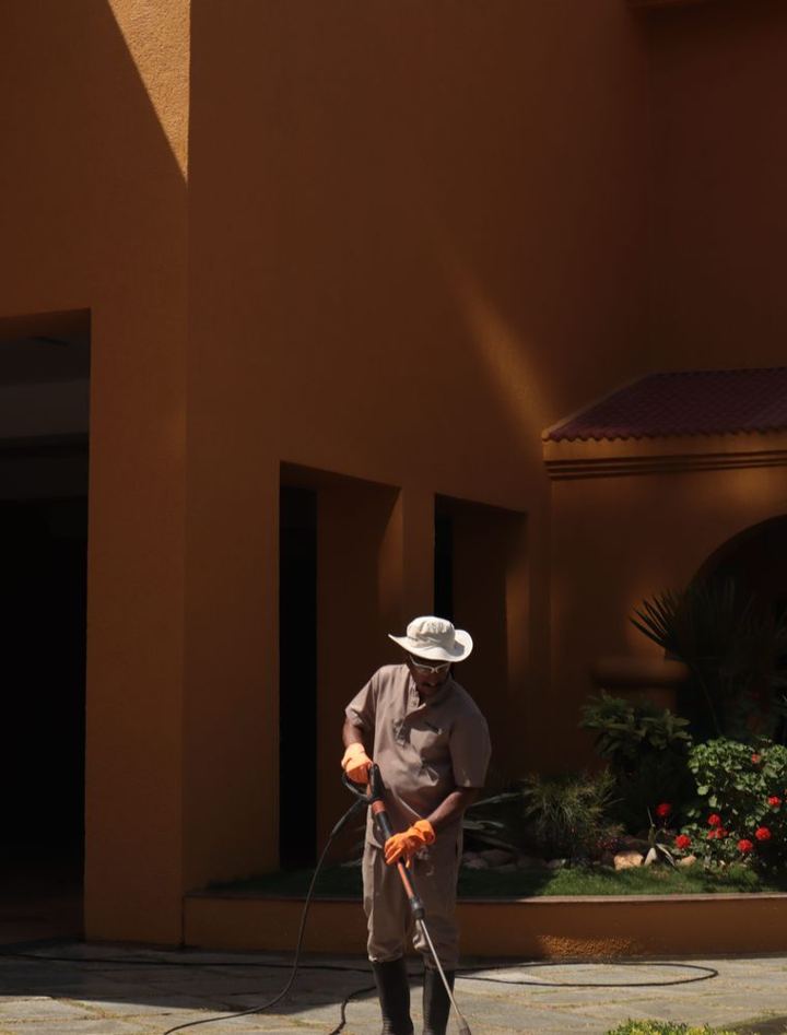 A worker in a wide-brimmed hat washing the walkway beside a stucco house